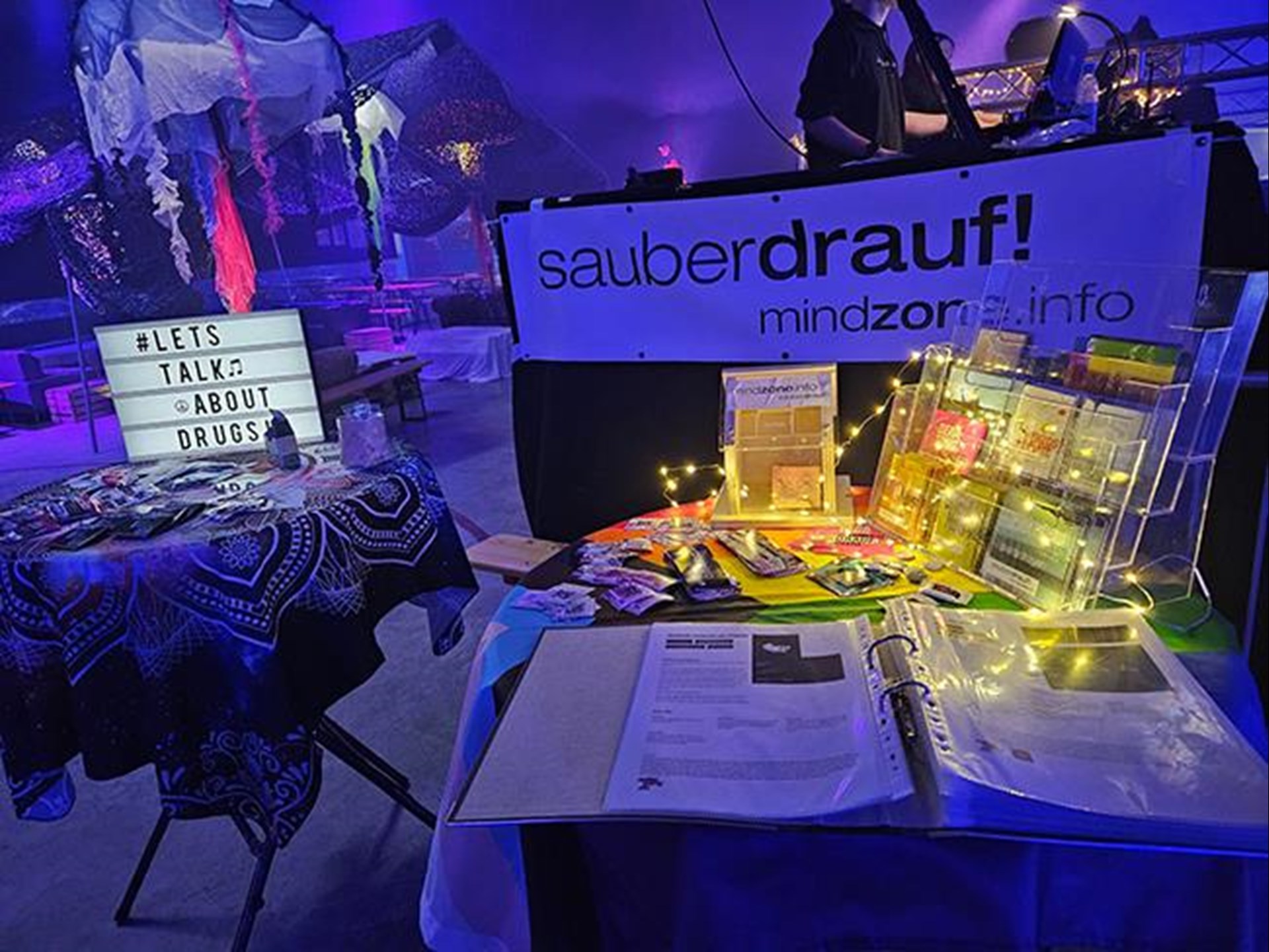 In violett dunkelblaues Licht ist der Info-Stand von Mindzone bei Syrens Festival eingetaucht. Man sieht im Vordergrund die Vielzahl von Info-Materialien. Im Hintergrund steht ein Musiker der Band. 
