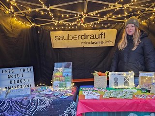 Unter dem Motto "Sauber drauf" warb Mindzone Augsburg auf der "Techno Party mit Punsch" für Aufklärung über Drogen. Eine Lichterkette beleuchtet den Infostand. Im Vordergrund sieht man die vielen Info-Materialien. Die Leitung von Mindzone ist ebenfalls zu sehen. Sie trägt eine Mütze, um sich vor der Kälte zu schützen. 